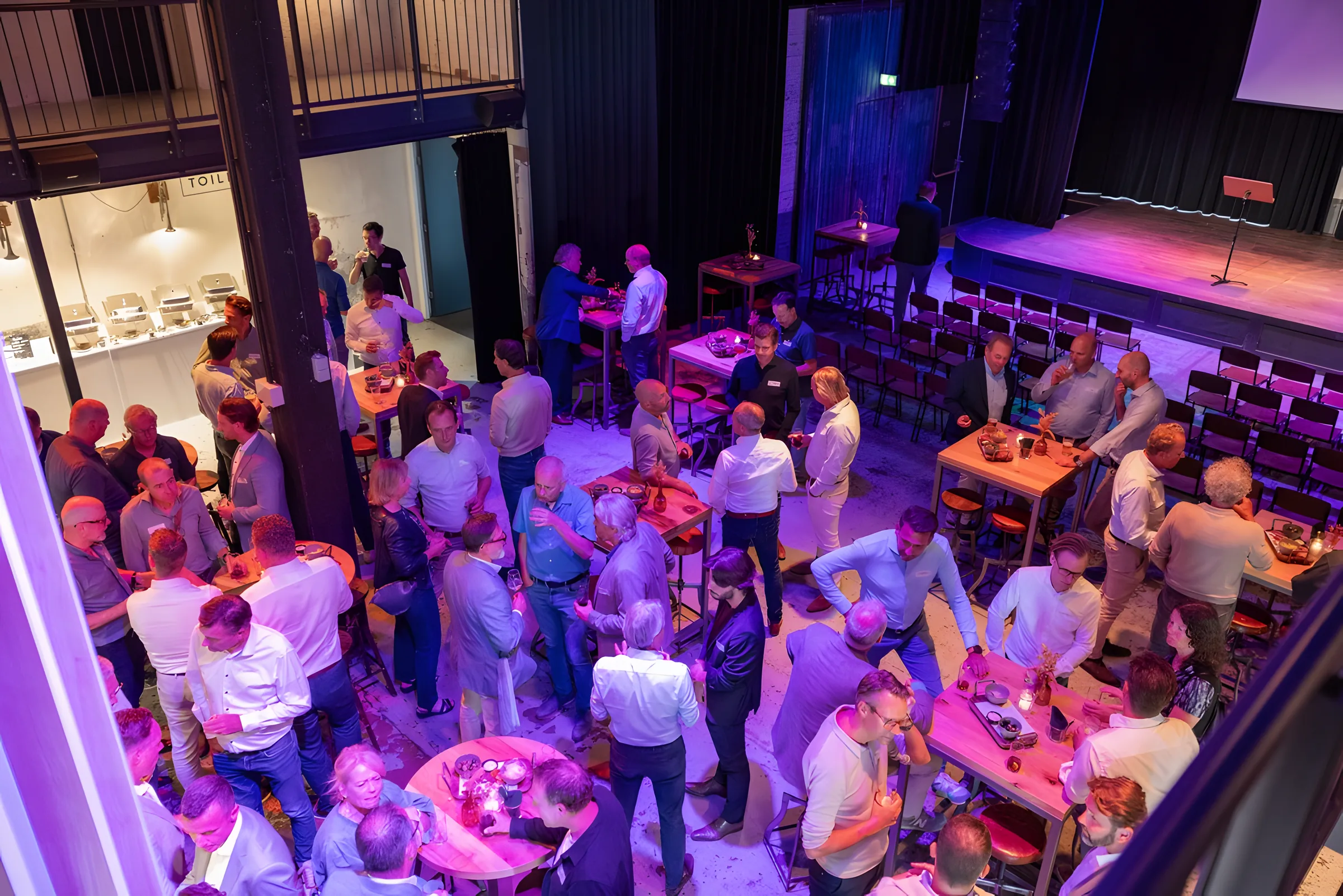 Leden van de Bouwsociëteit in gesprek aan de statafels tijdens een avondbijeenkomst, van bovenaf